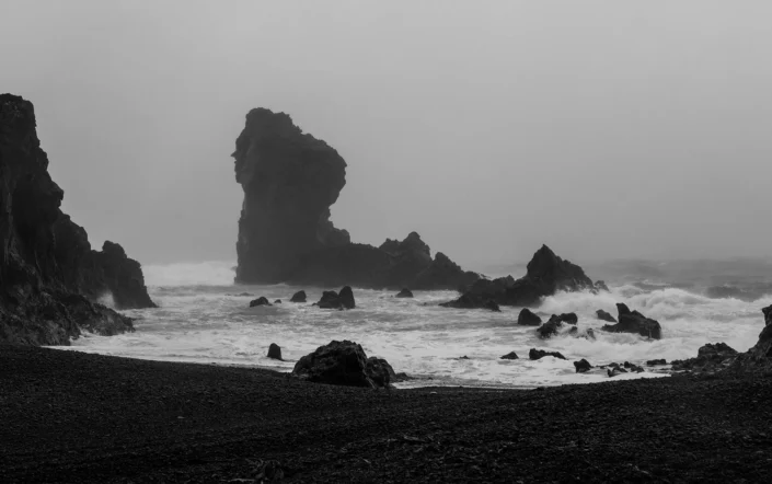 Photo en noir et blanc d'un rocher sur une plage volcanique au milieu de la tempête en Islande